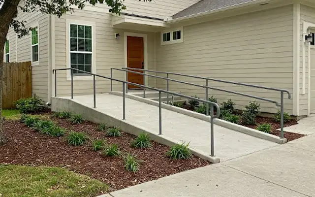 Concrete wheelchair ramp with metal handrails and mulched planting beds at a home entrance