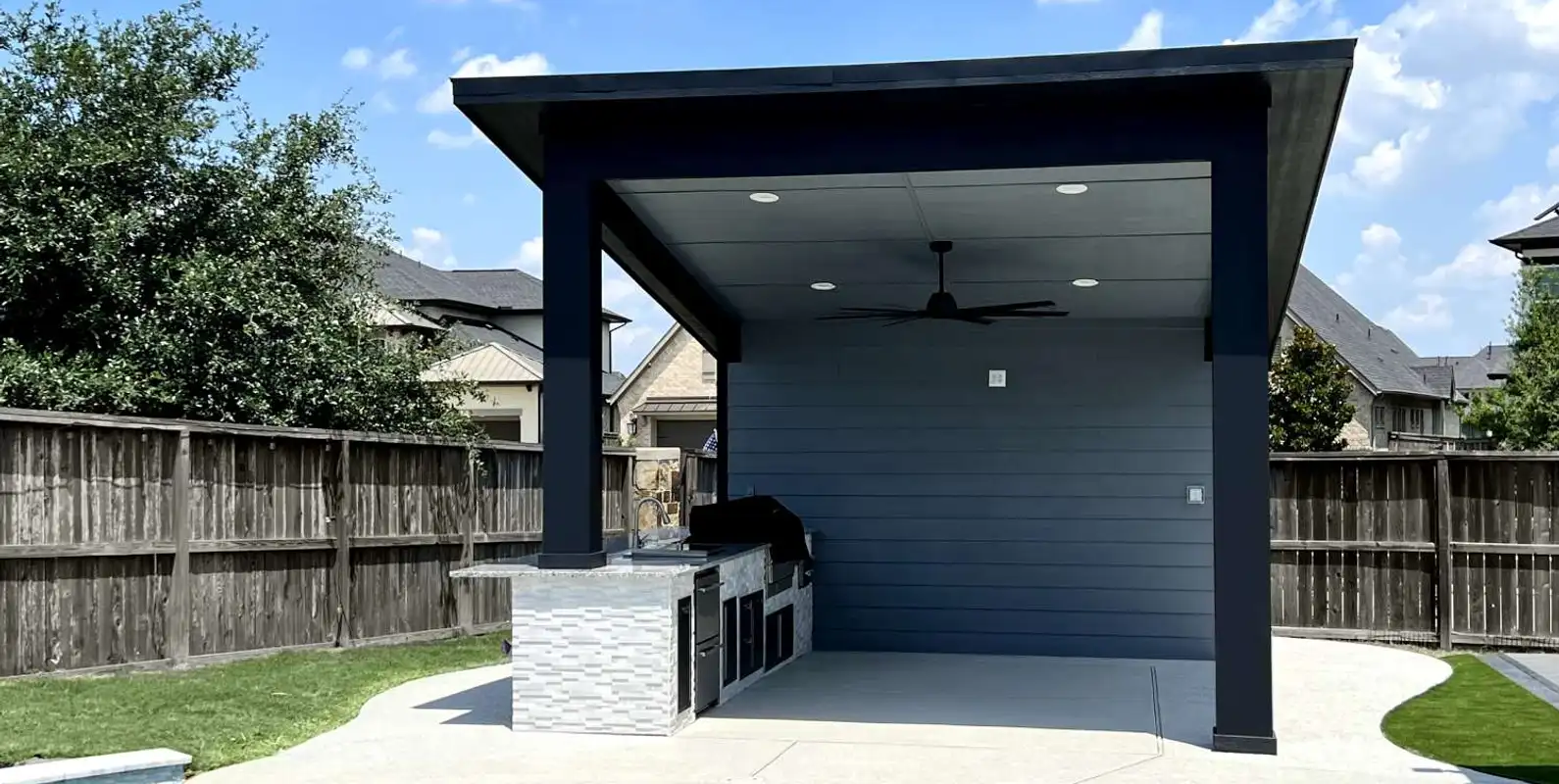 Modern covered pavilion with ceiling fan and stone-clad outdoor kitchen beside a backyard pool