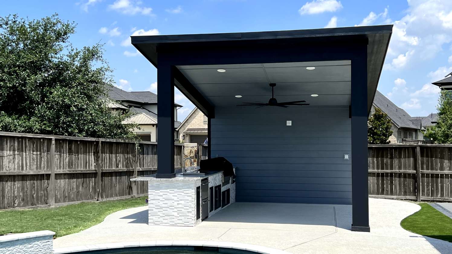 Modern covered pavilion with ceiling fan and stone-clad outdoor kitchen beside a backyard pool