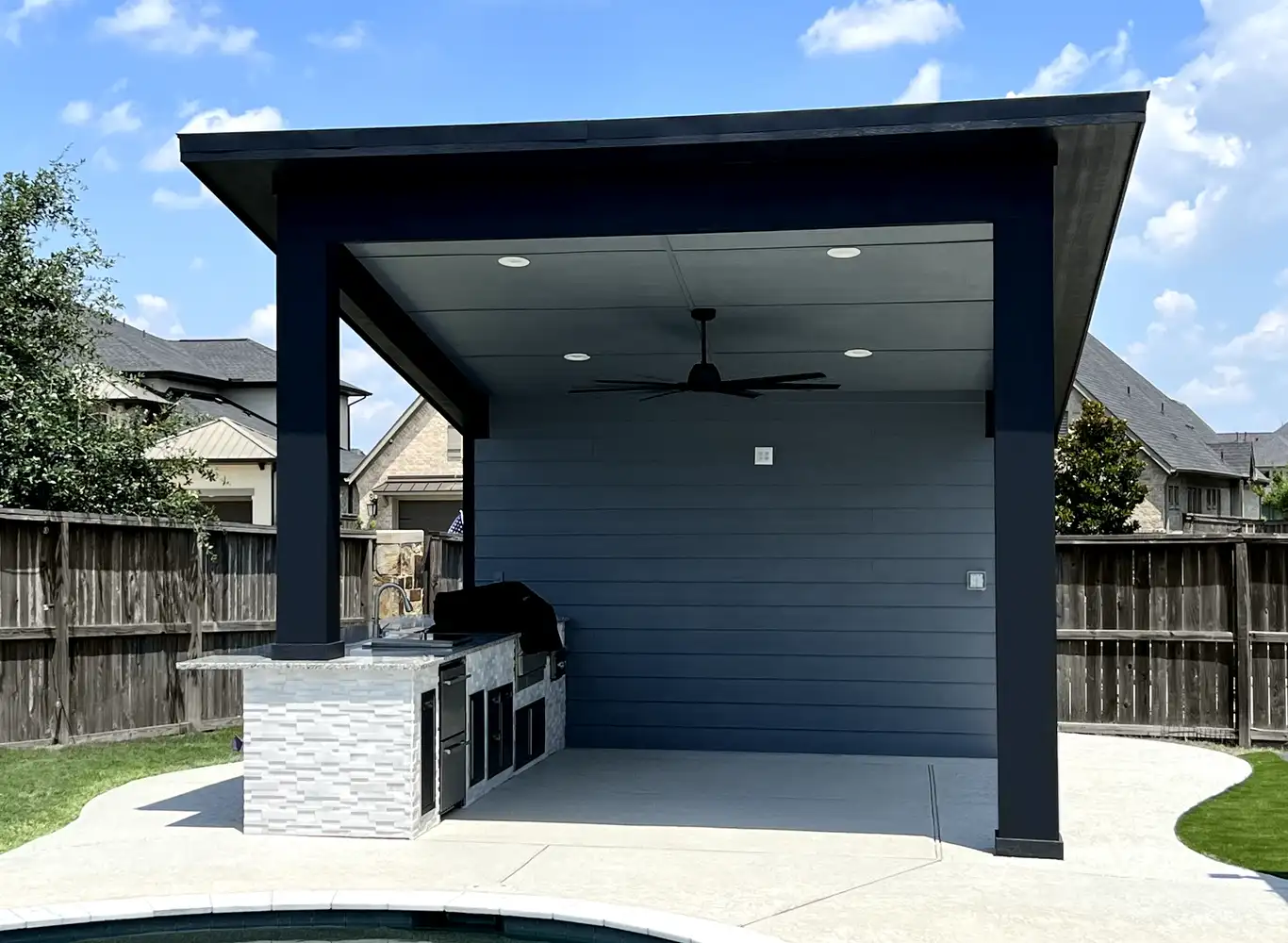 After: extended pool deck with a modern covered pavilion, ceiling fan and stone-clad outdoor kitchen