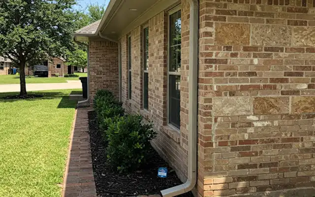 Gutter downspout on a brick house beside a mulched shrub bed with brick paver edging
