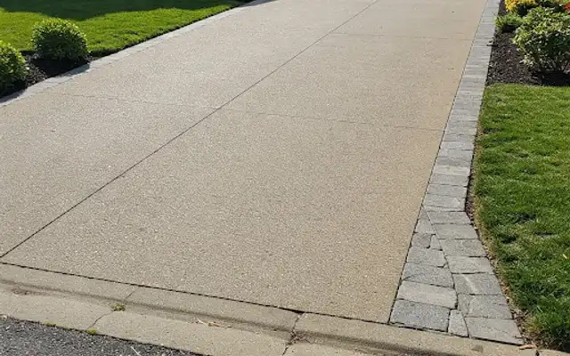 Wide broom-finish concrete driveway with a gray paver border and flower beds