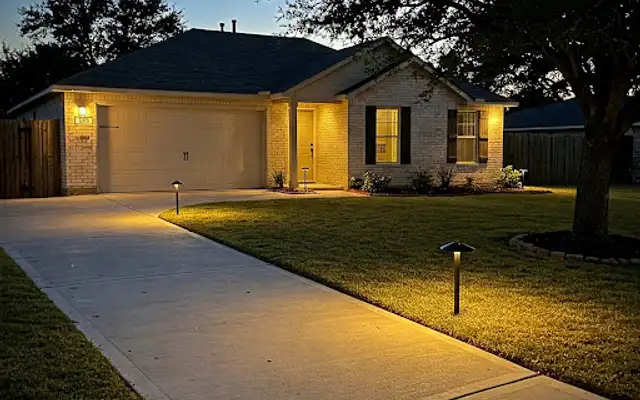 Concrete driveway at dusk lined with low-voltage path lights beside a brick home