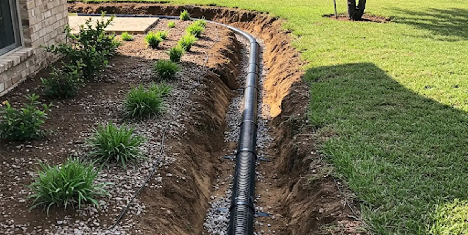 Open trench with corrugated drain pipe laid on gravel beside a lawn and planting bed