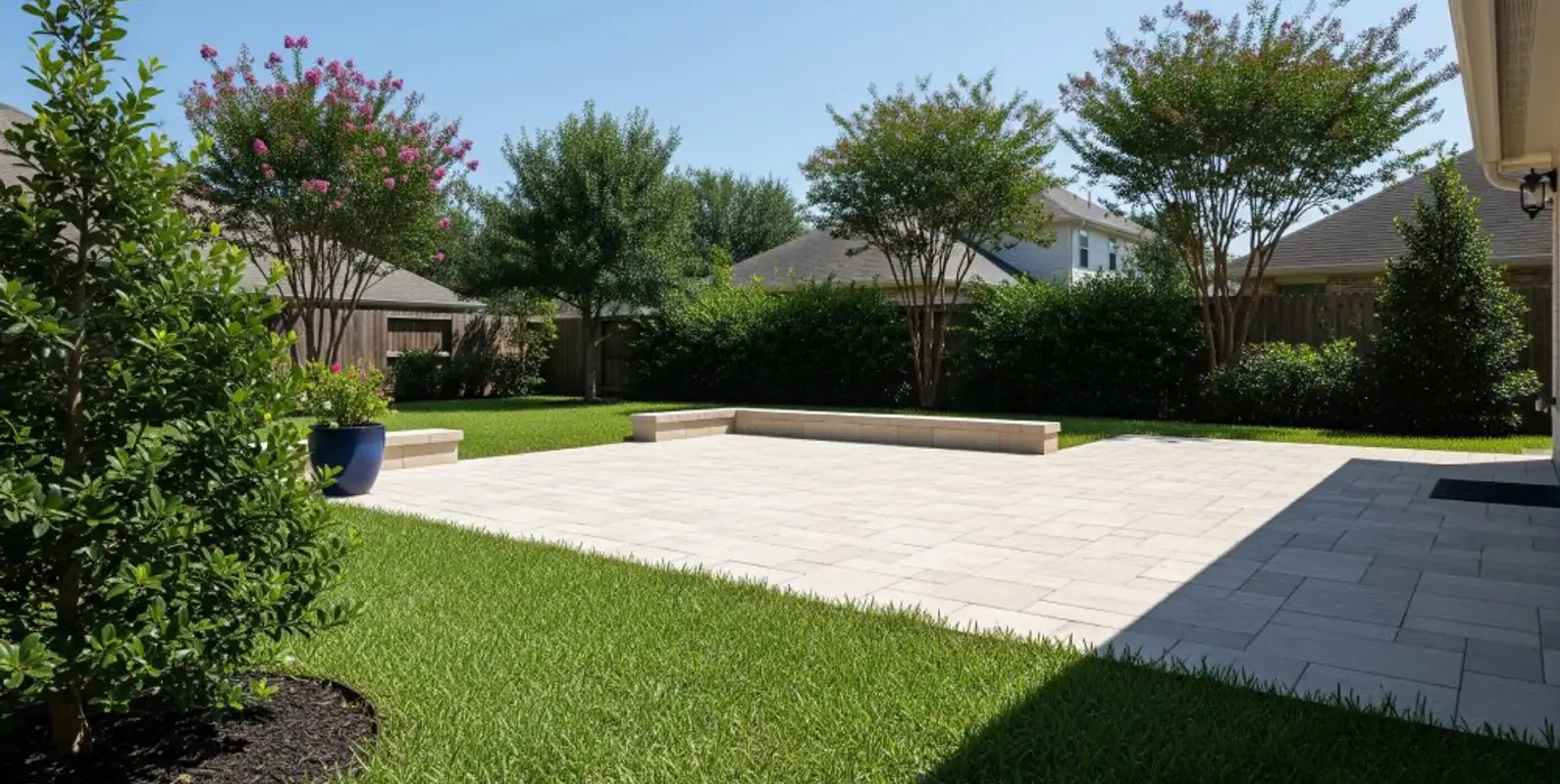 Light tan paver patio surrounded by green lawn and crape myrtles in a backyard