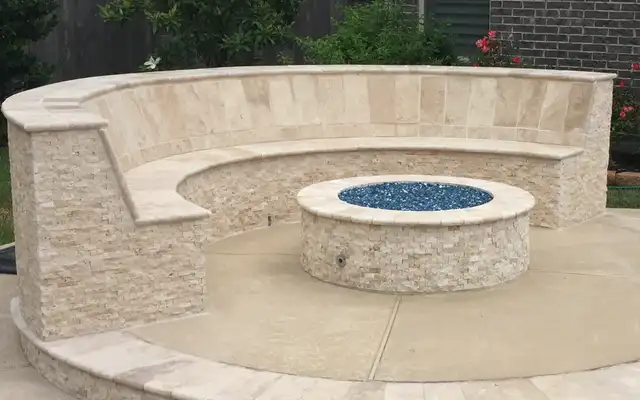 Round split-face stone fire pit with blue fire glass inside a curved stone seat wall by a pool, built by HLI