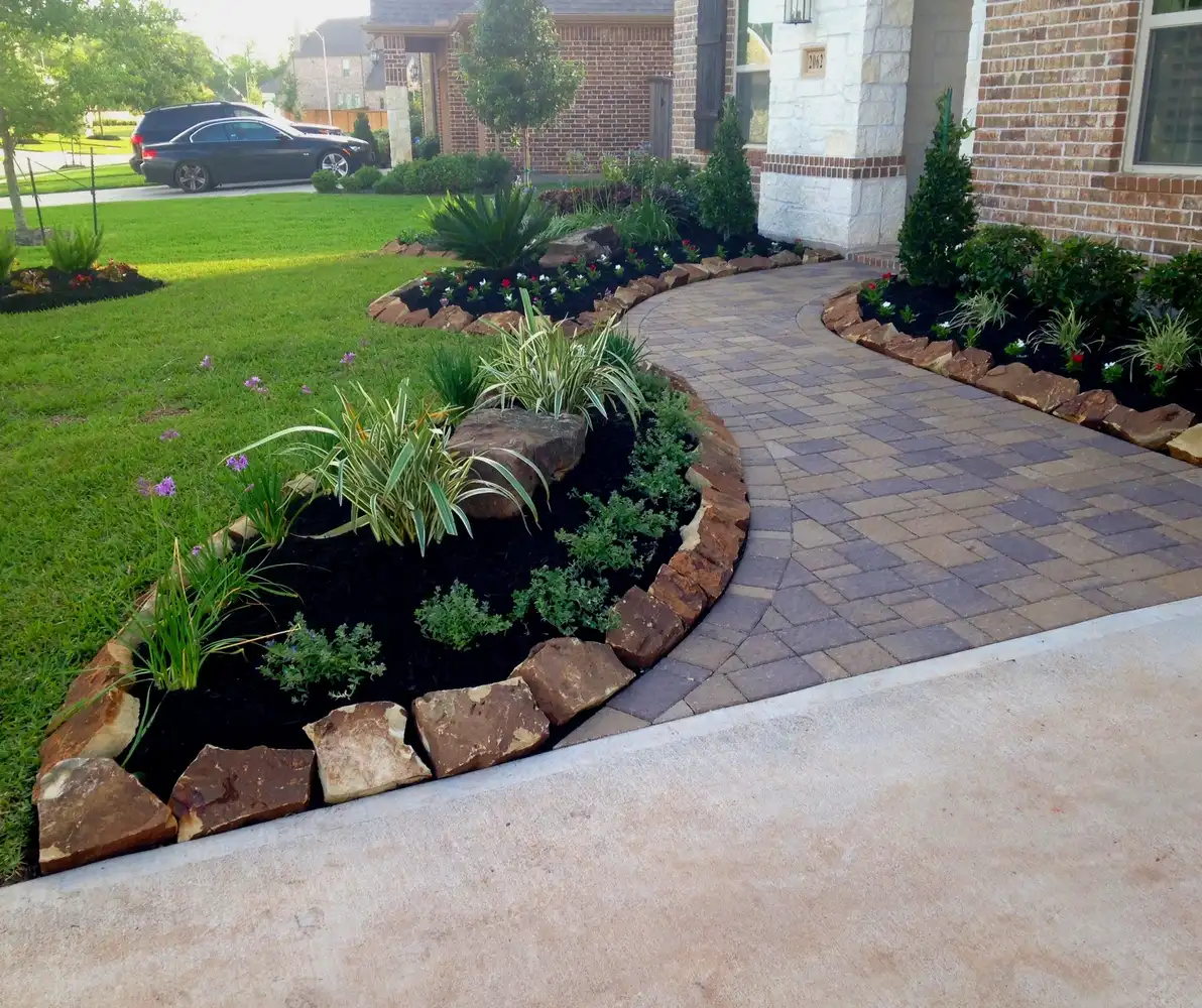 Curved paver walkway with boulder-edged mulched beds and ornamental grasses at a brick home, built by HLI