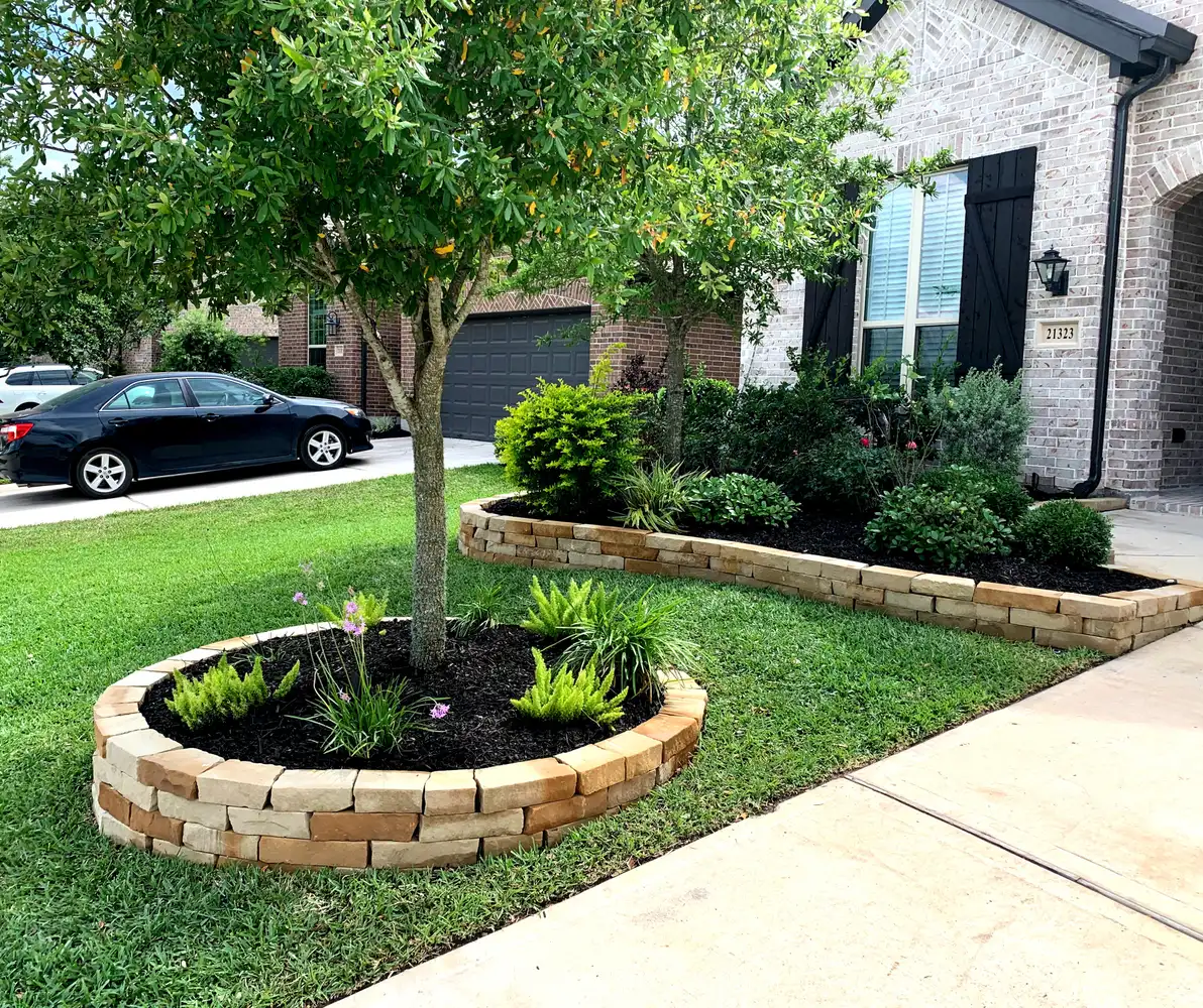 Stacked stone tree ring and raised stone-bordered shrub bed in a green front lawn, built by HLI