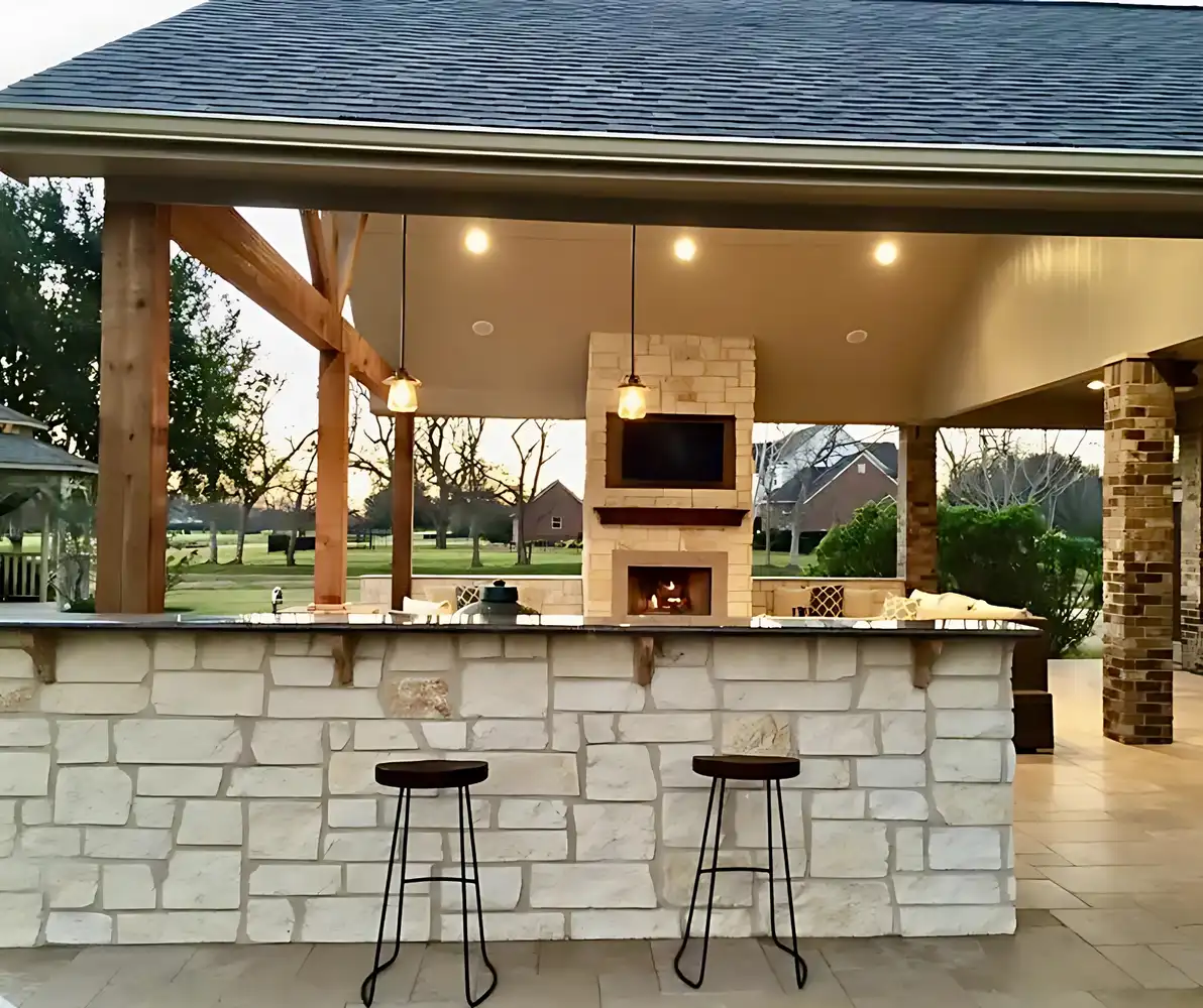 Limestone outdoor kitchen bar with pendant lights, fireplace and TV under a patio cover at dusk, built by HLI