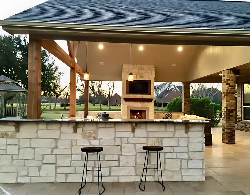 Limestone outdoor kitchen bar with pendant lights, fireplace and TV under a patio cover at dusk
