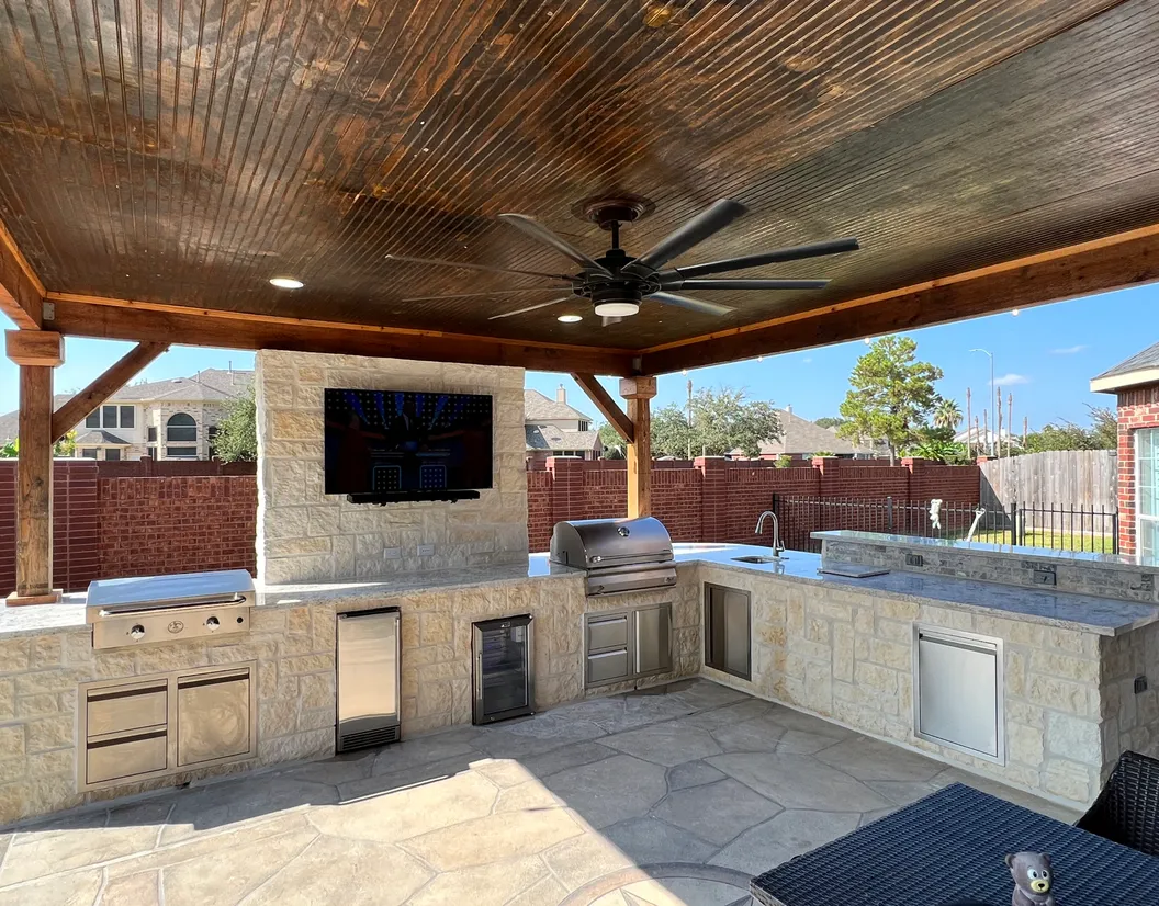 Limestone outdoor kitchen with grill, sink and TV wall under a stained cedar patio cover