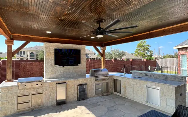Limestone outdoor kitchen with grill, sink and TV wall under a stained wood ceiling