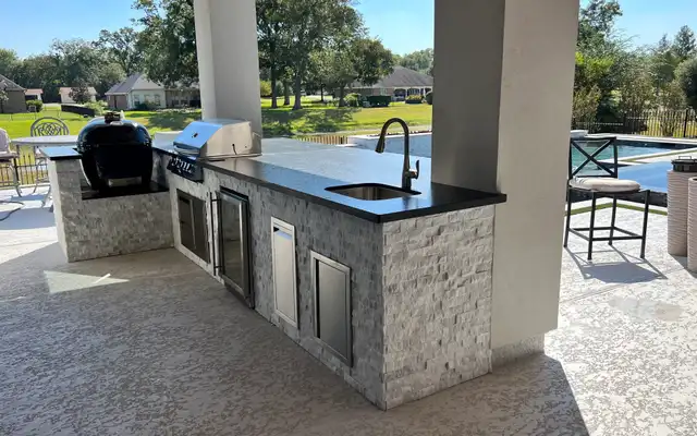 Split-face stone kitchen island with kamado, grill and sink beside a pool