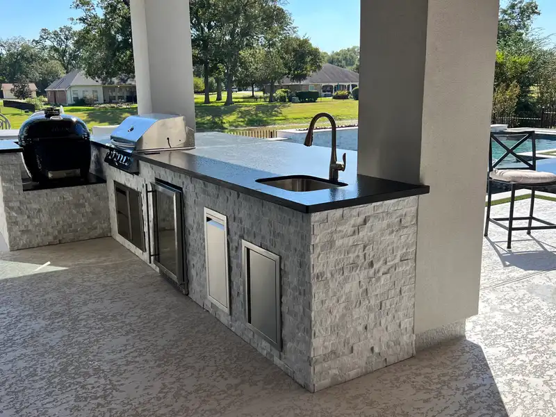Stacked stone outdoor kitchen island with grill, kamado cooker and sink overlooking a pool