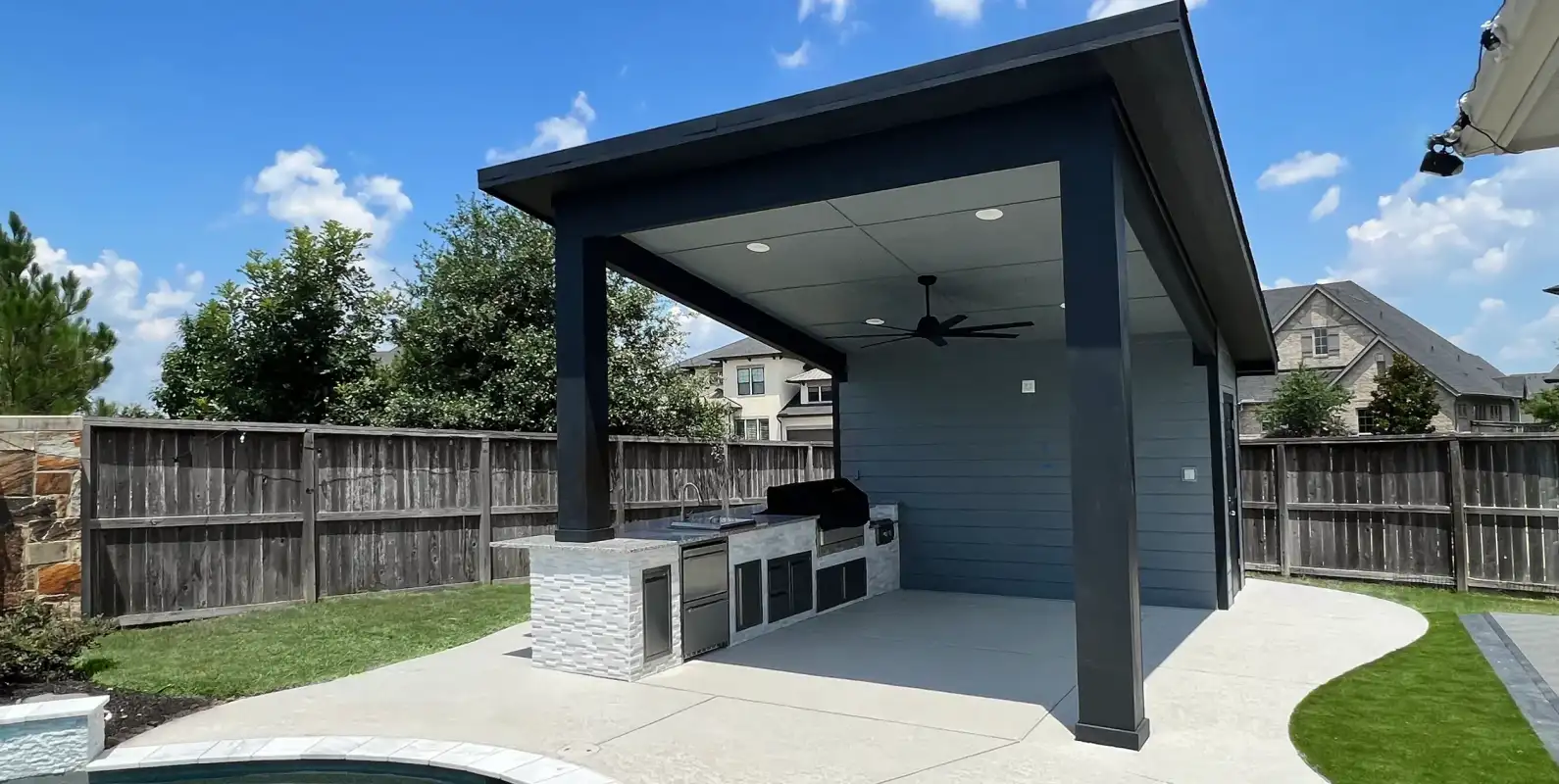 Modern dark pavilion with outdoor kitchen and ceiling fan beside a backyard pool