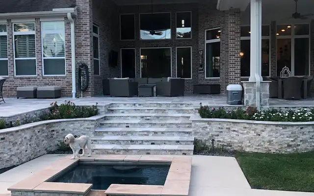 Travertine steps and stacked stone planter walls leading from a covered patio to a pool spa