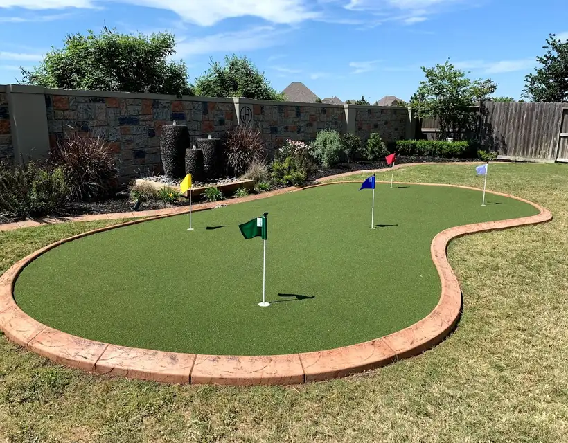 Artificial putting green with flags and stamped concrete curb edging near a stone wall