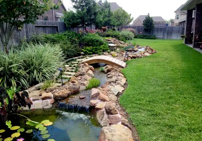 Rock-lined backyard stream with waterfall, arched wooden bridge and lily pond in a Houston yard