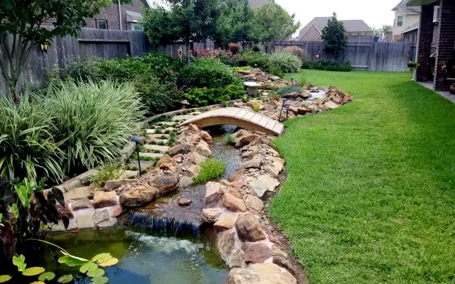Lily pond fed by a rock-lined stream and waterfall under an arched wooden bridge, built by HLI