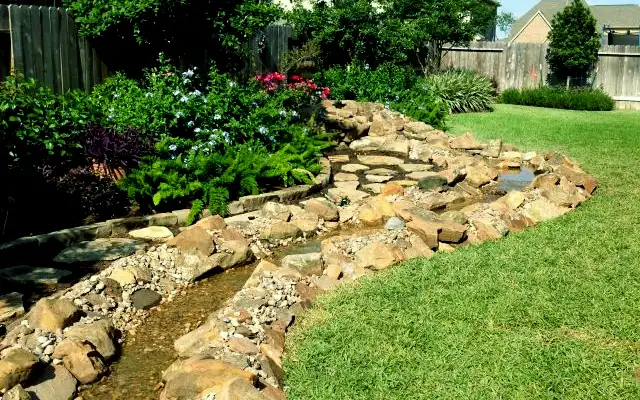 Boulder-lined stream bed with gravel winding along a lawn beside flowering shrubs and ferns, built by HLI