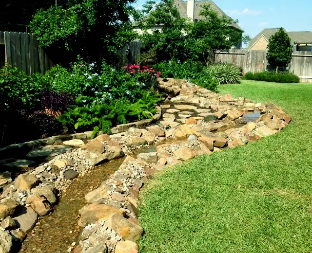 Boulder-lined dry creek bed winding along a lawn with flowering shrubs and ferns