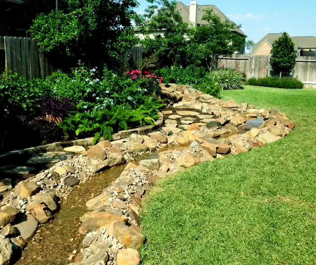 Boulder-lined backyard stream winding past a lawn, flowering shrubs and ferns, built by HLI