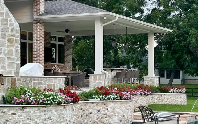 Covered patio with stacked stone planter walls full of red and pink flowers beside a fire pit edge