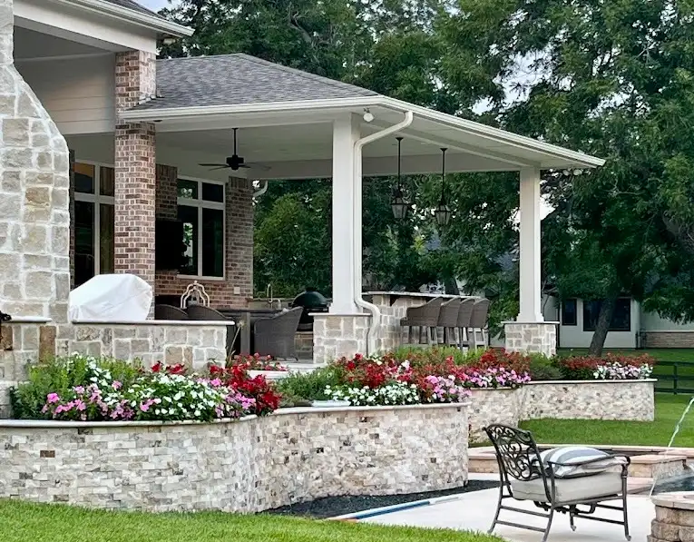 Stacked stone planter walls planted with red and pink flowers beside a covered patio