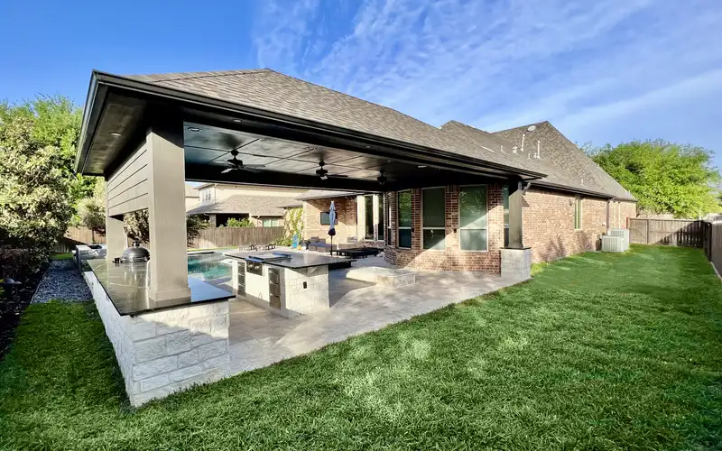 Covered patio with stone outdoor kitchen island overlooking a pool and Houston backyard lawn