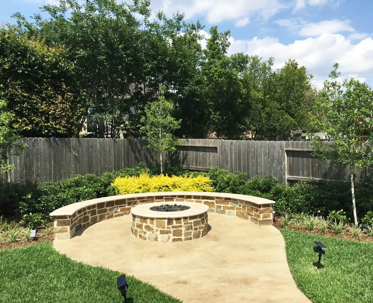 Round flagstone fire pit with a curved seat wall, shrubs and landscape lights on a concrete patio