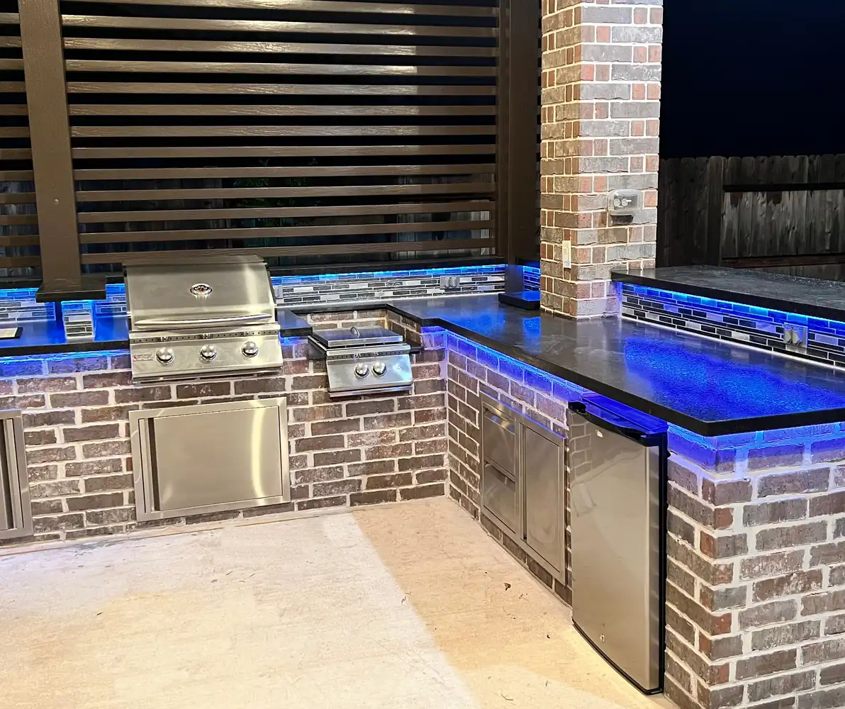 Brick outdoor kitchen with blue LED under-counter lighting and stainless grill at night