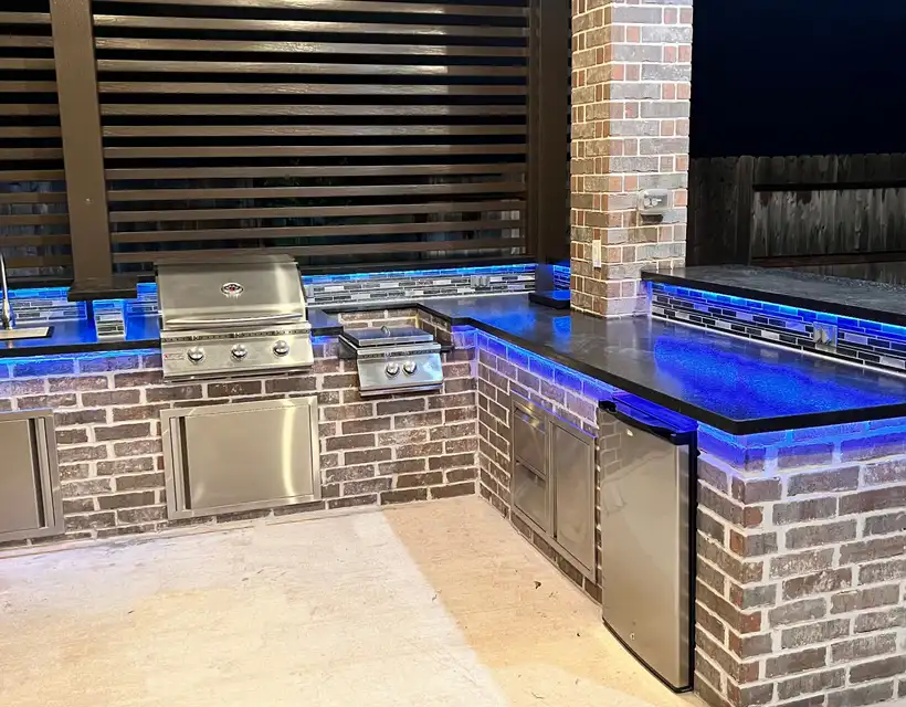 Brick outdoor kitchen with blue LED under-counter lighting and a slat screen at night