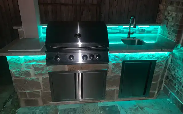 Stone outdoor grill station with teal LED under-counter lights and a sink at night