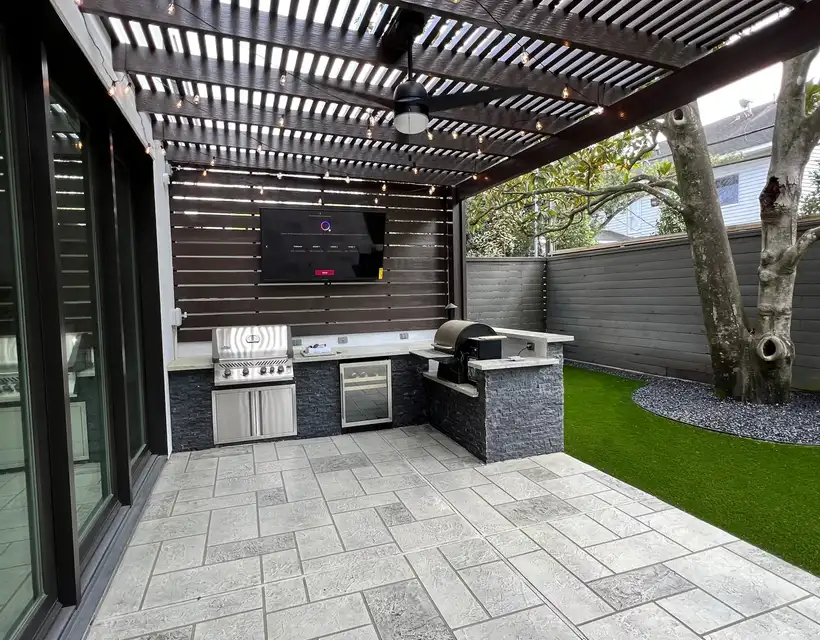 Modern stone outdoor kitchen under a dark slatted pergola beside a live oak