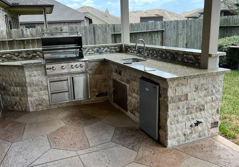 L-shaped stone outdoor kitchen with grill, sink and mini fridge on stamped concrete