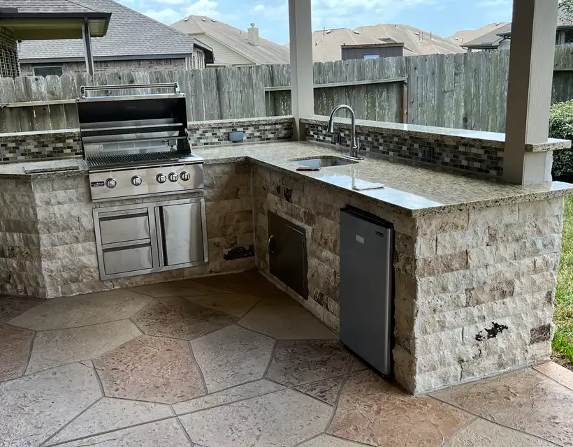 L-shaped stone outdoor kitchen with grill, sink and mini fridge on stamped concrete