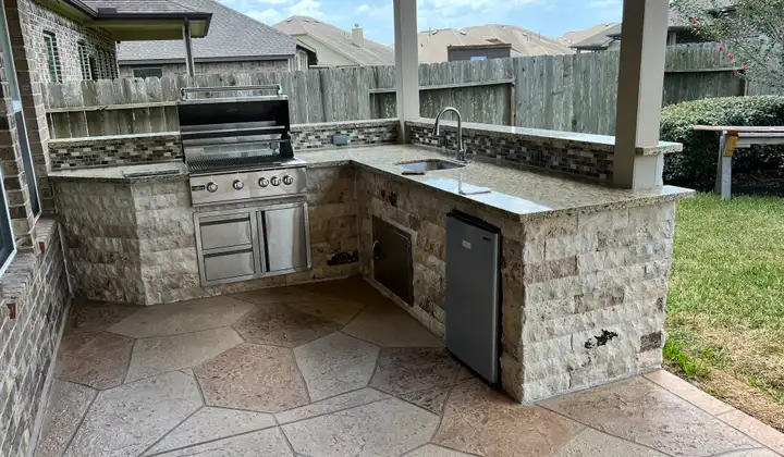 L-shaped stone outdoor kitchen with grill, sink and mini fridge on stamped concrete