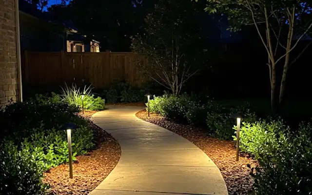 Curved concrete walkway at night lined with low-voltage path lights and shrubs