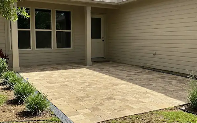 Tan paver patio with a channel drain running along its edge behind a beige house