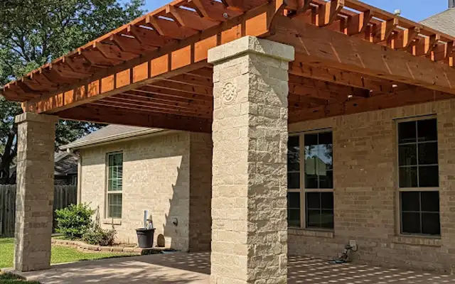 Stained cedar pergola on stone-clad columns over a patio behind a brick home