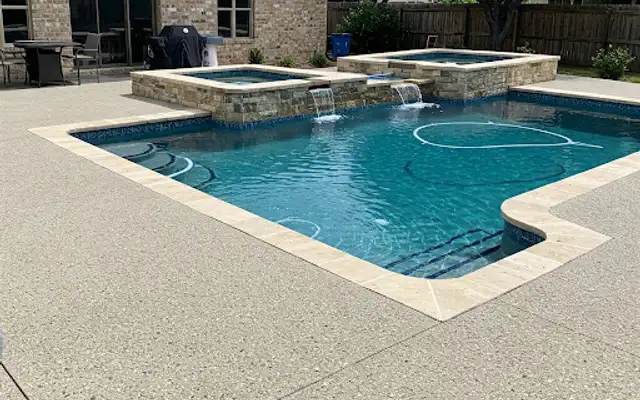 Textured concrete pool deck around a pool with a raised stone spa and spillways