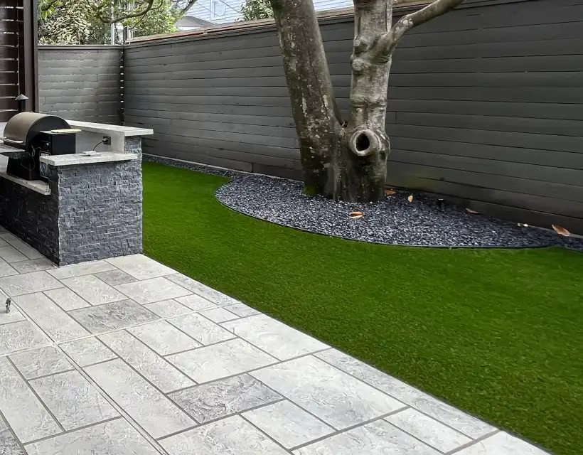 Artificial turf between large gray pavers, a black rock tree ring and an outdoor grill island