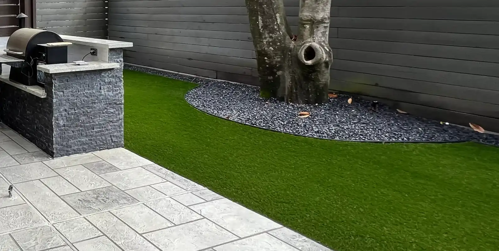 Artificial turf lawn beside large gray pavers and a black rock tree ring in a backyard